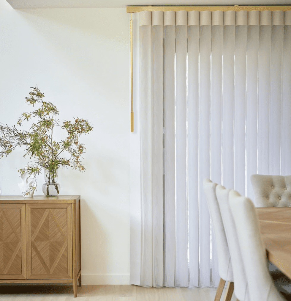 A dining area with a wooden table and upholstered chairs, a sideboard with a glass vase holding branches, and white vertical blinds covering a window.