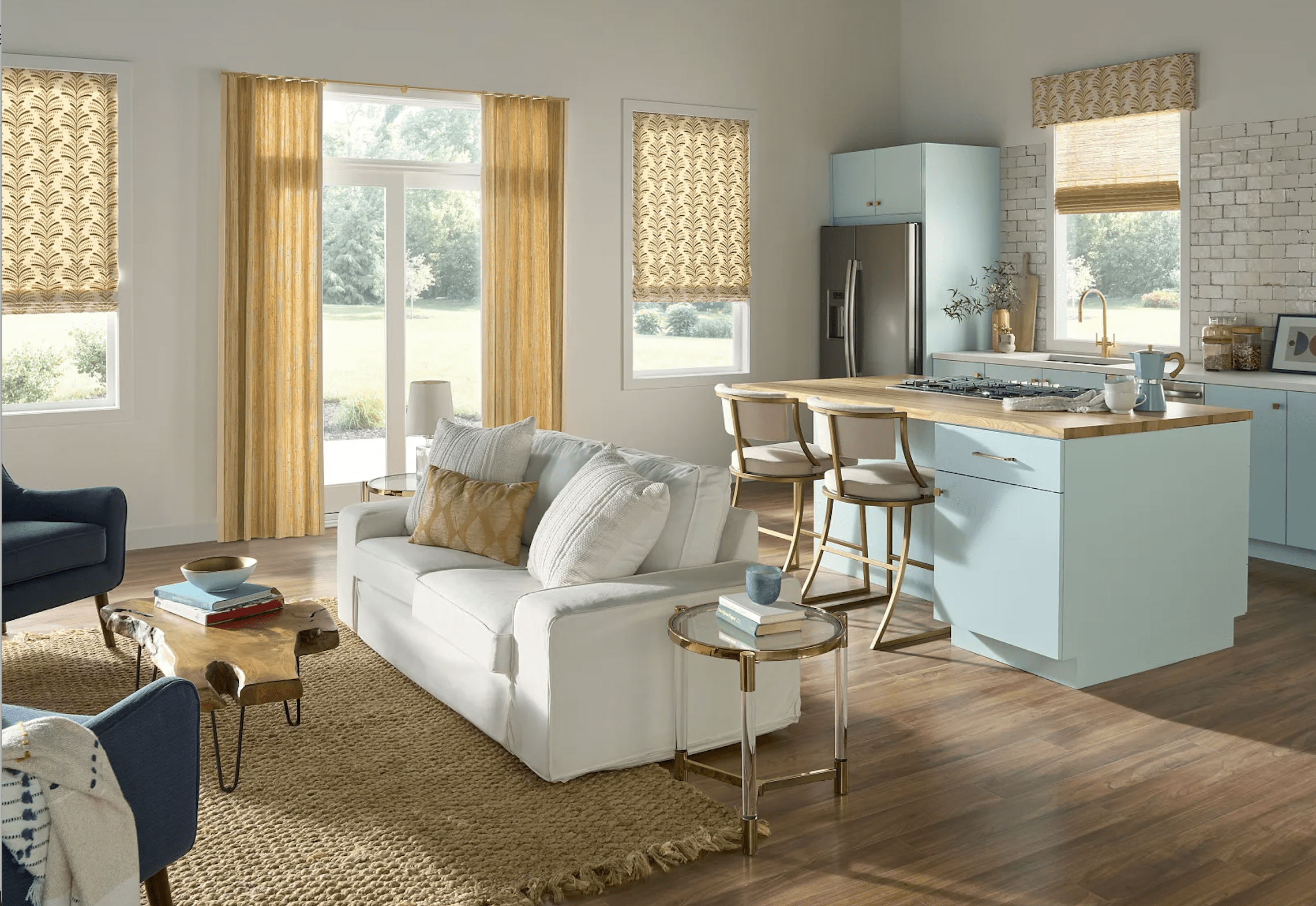 Open-concept living room and kitchen with light blue cabinets, wood countertops, a white sofa, woven rugs, bar stools, and large windows with patterned curtains.