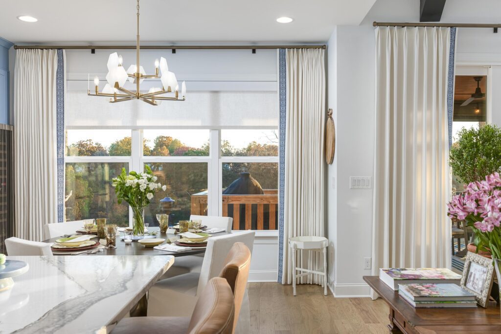 A bright dining area with a marble table, white chairs, flowers, and large windows with white curtains overlooking an outdoor deck and trees.