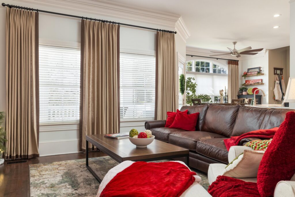 A living room with a brown leather sectional sofa, red and white cushions and blankets, a wooden coffee table, large windows with beige curtains, and a ceiling fan in the background.