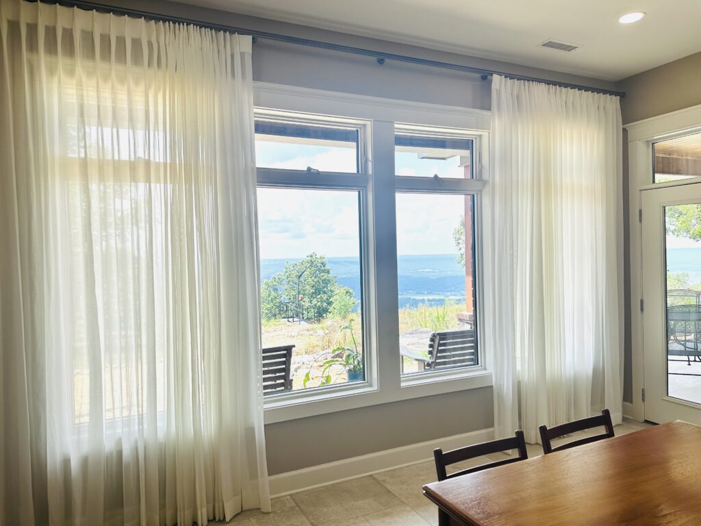 Large windows with sheer white curtains reveal a scenic view of water, trees, and sky from an indoor dining area with a wooden table and chairs.