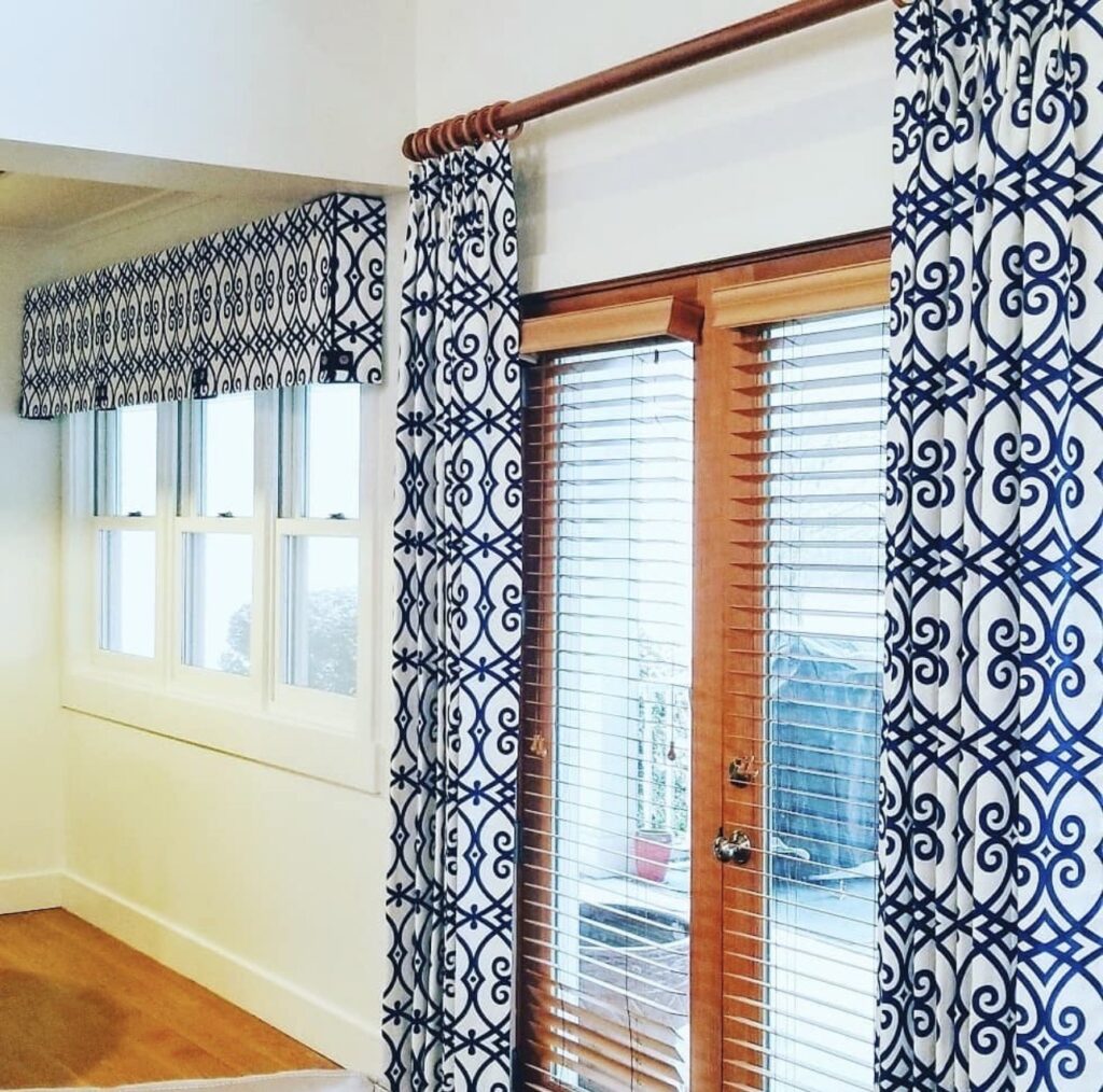 French doors with wooden blinds and blue patterned curtains next to a set of windows with a matching blue patterned valance in a bright room with white walls.