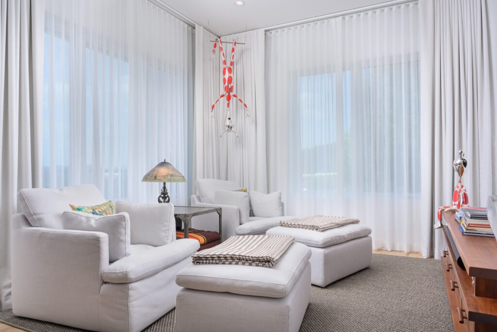 Bright living room with white armchairs, ottomans, sheer curtains, a side table with a lamp, and red-and-white hanging decor. Light fills the space through large windows.