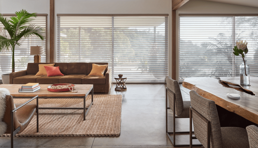 Modern living room with a brown sofa, colorful pillows, wooden coffee table, woven rug, large windows with blinds, and a dining area with a wooden table and chairs.