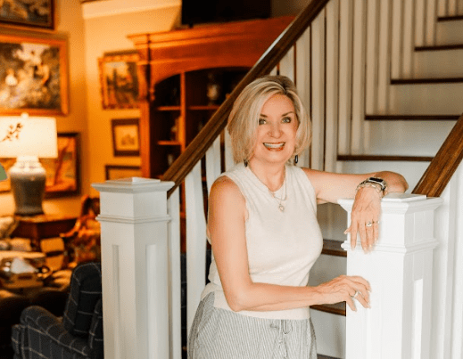 A woman with short blonde hair, wearing a sleeveless top and striped pants, stands smiling by a staircase in a warmly decorated living room.