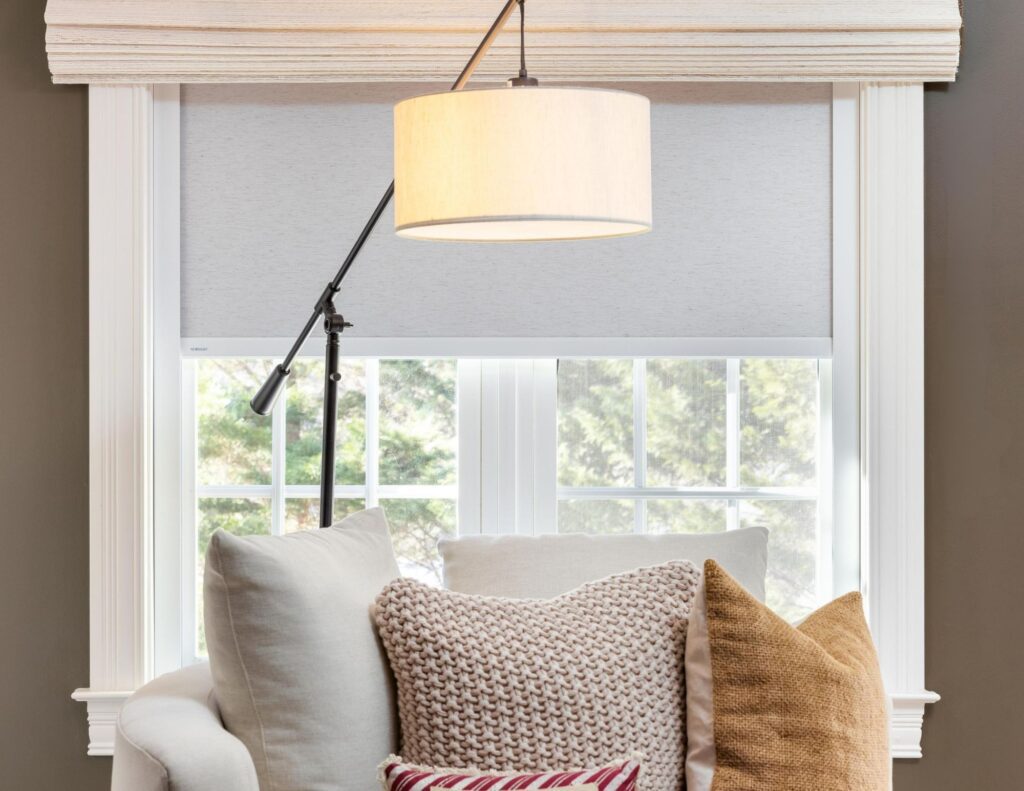 A white armchair with three textured pillows sits in front of a window with white blinds and a cream-colored lamp hanging above.
