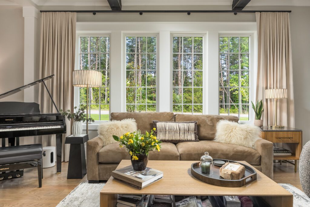 A living room with a brown sofa, a coffee table with flowers, large windows with beige curtains, a piano, and a view of greenery outside.
