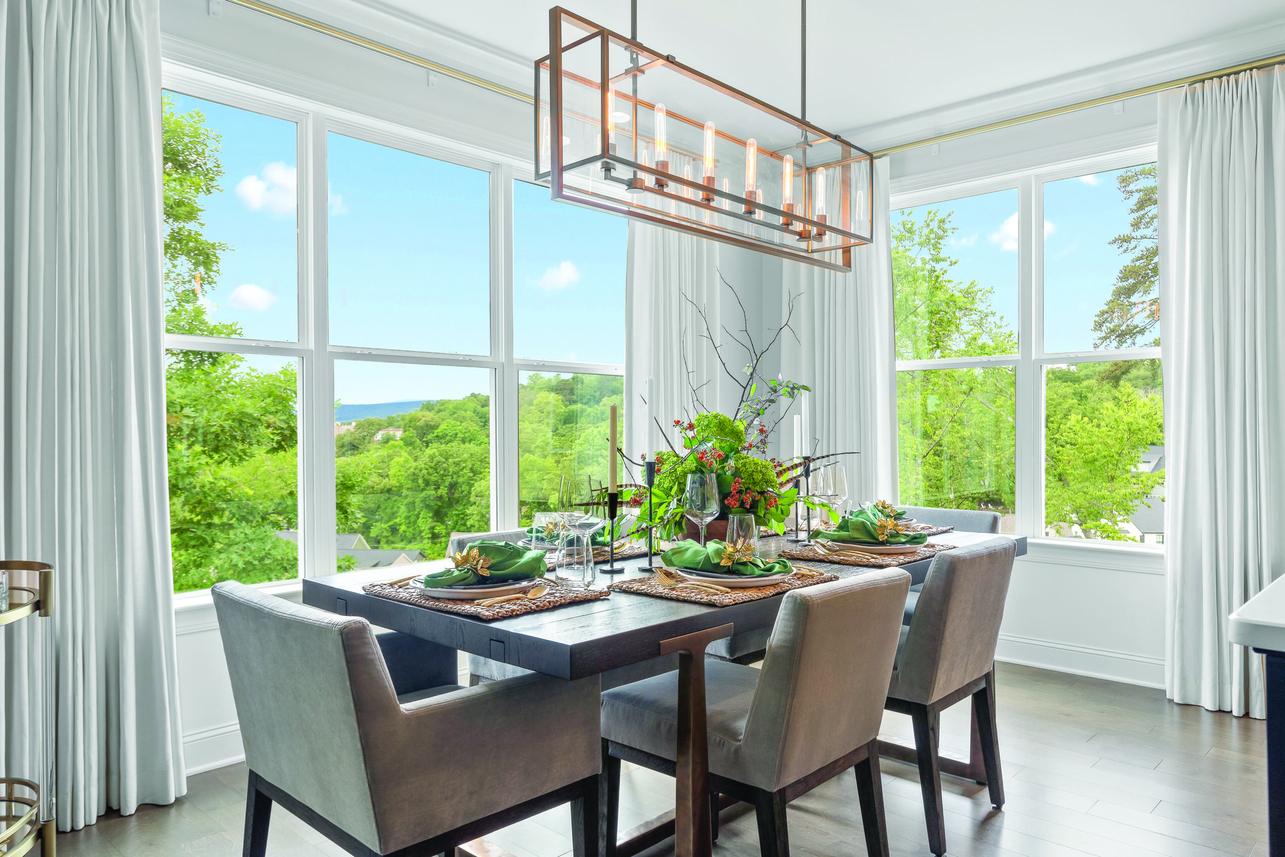 Modern dining room with a dark wooden table set for four, gray upholstered chairs, large windows with white curtains, and a rectangular chandelier overhead. Lush greenery is visible outside.