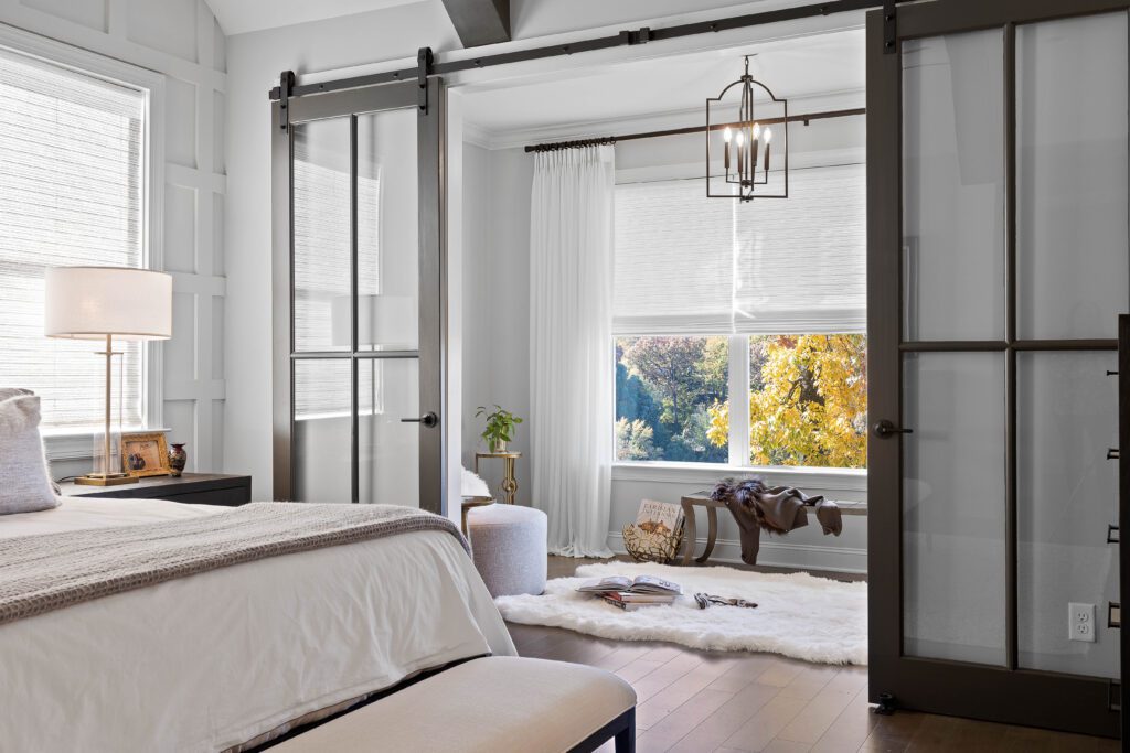 Modern bedroom with glass sliding doors opening to a sunlit sitting area, featuring a large window, white rug, and contemporary decor.