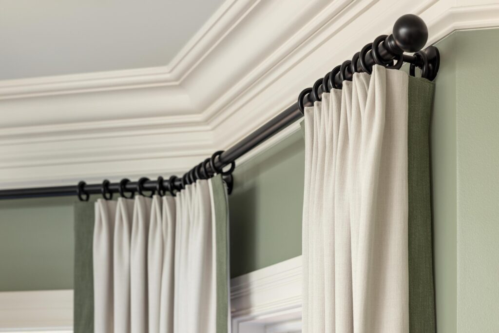 A close-up view of beige curtains with dark green side panels, hanging from a black curtain rod in a corner with crown molding.