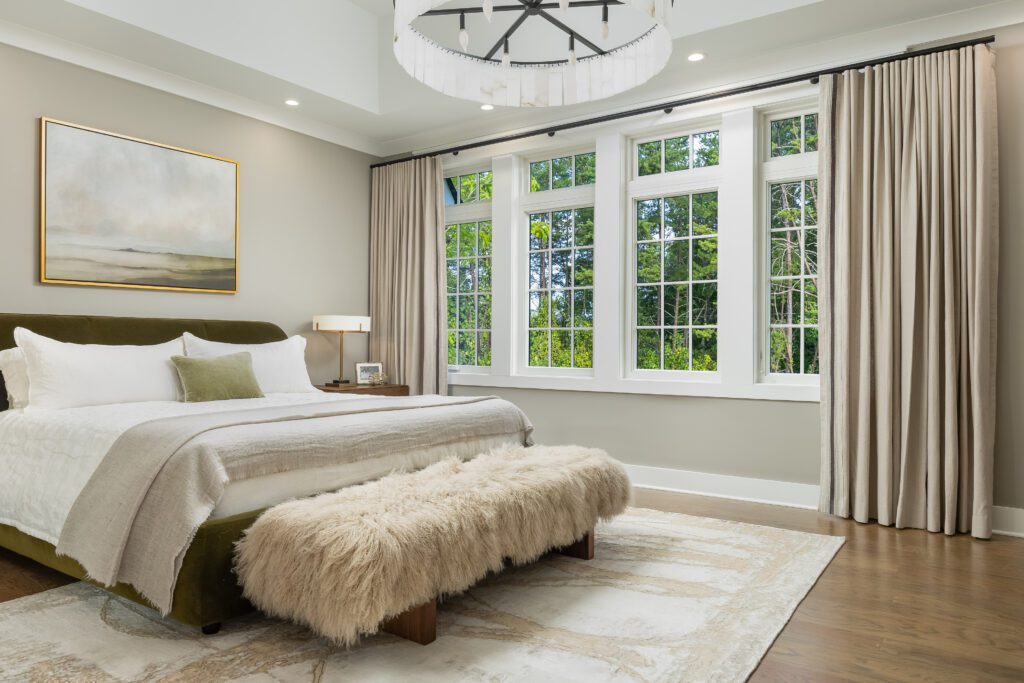 A modern bedroom with a large bed, a furry bench, a landscape painting, a rug, floor-length curtains, and tall windows overlooking greenery.