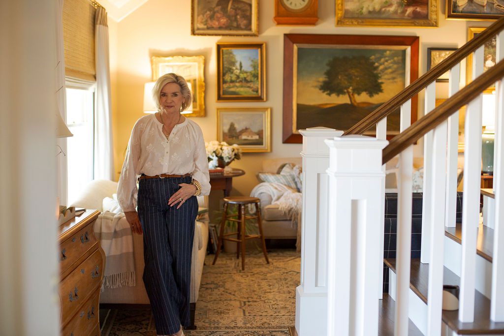 A woman stands in a living room decorated with framed paintings, a staircase, a couch, and wooden furniture visible in the background.