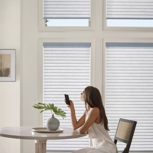 A woman in a white dress sits at a round table, holding a smartphone near her face. There is a potted plant on the table, and large windows with blinds in the background.